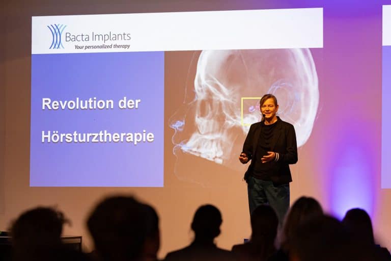 Dr. Verena Scheper standing on the stage pitching Bacta Implants on the SMINT Demo Day. Her pitch prersentation is seen in the background: The title is revolution in hearing rehabilitation